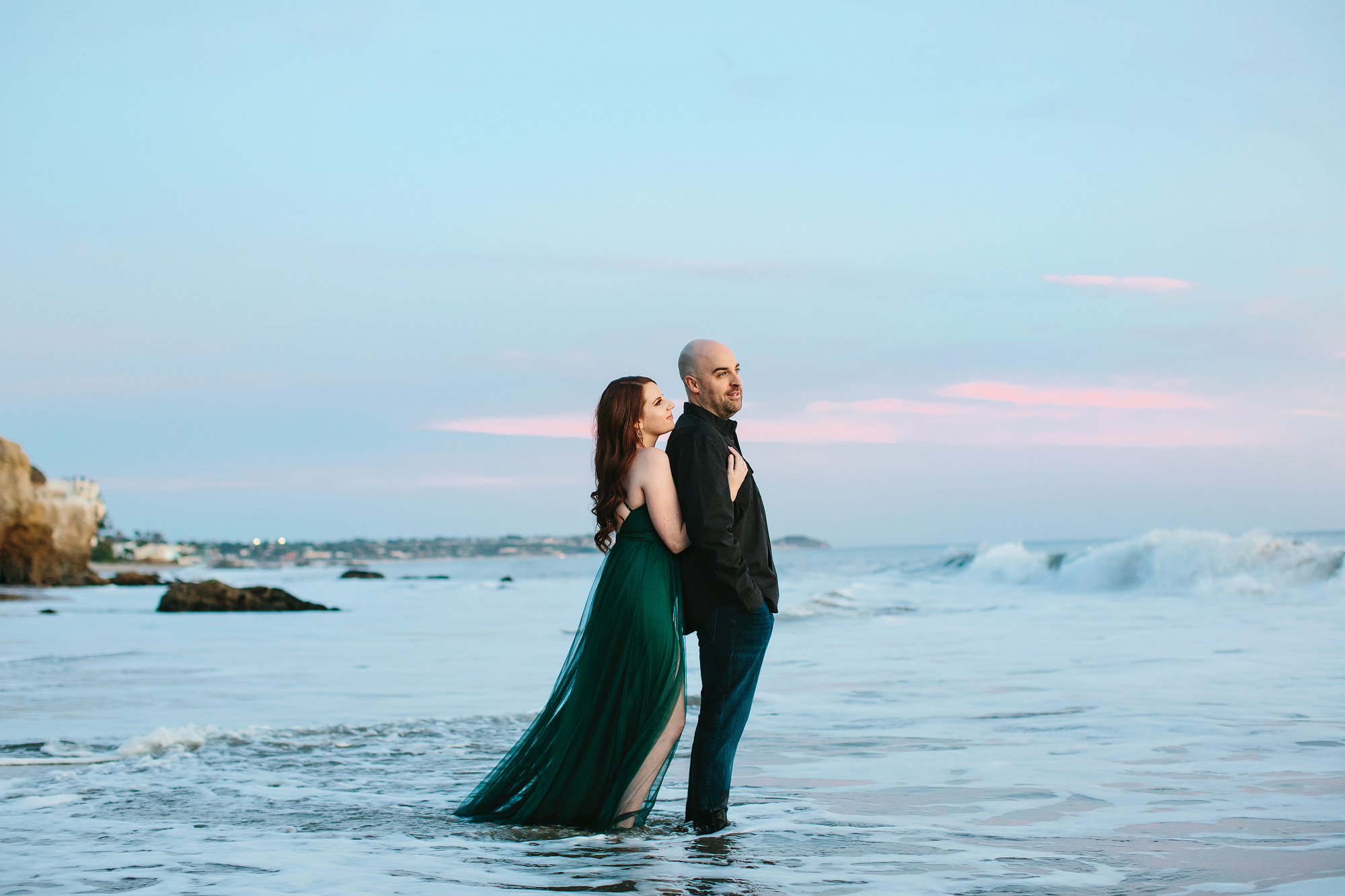 woman in green flowing dress hugging her fiance from behind while standing in the water at the beach in malibu california