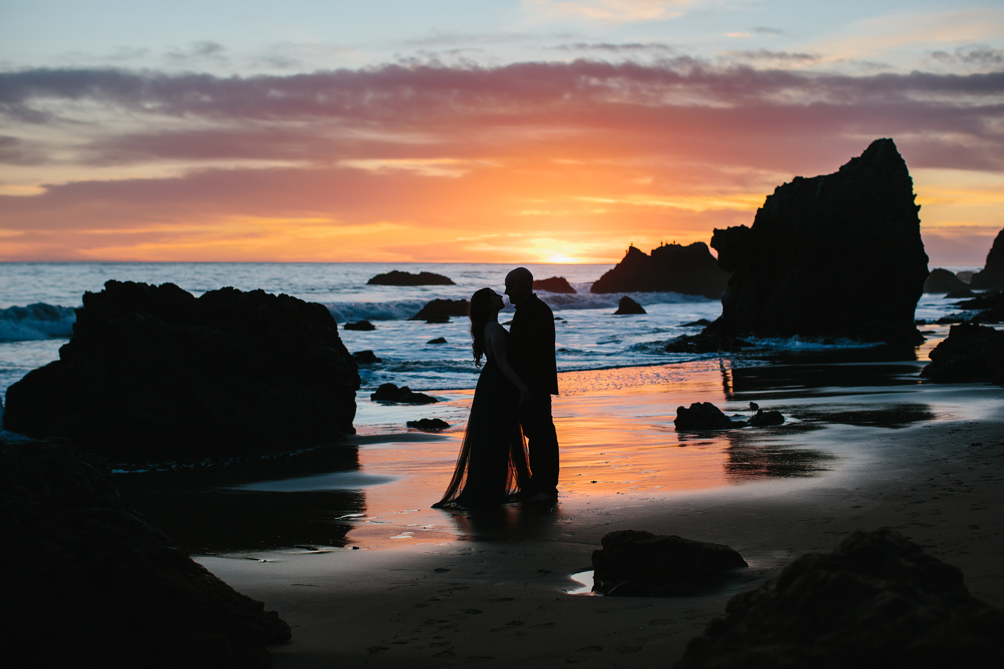 Silhouette of an engaged couple on the beach with the sunset behind them and the rocks of malibu