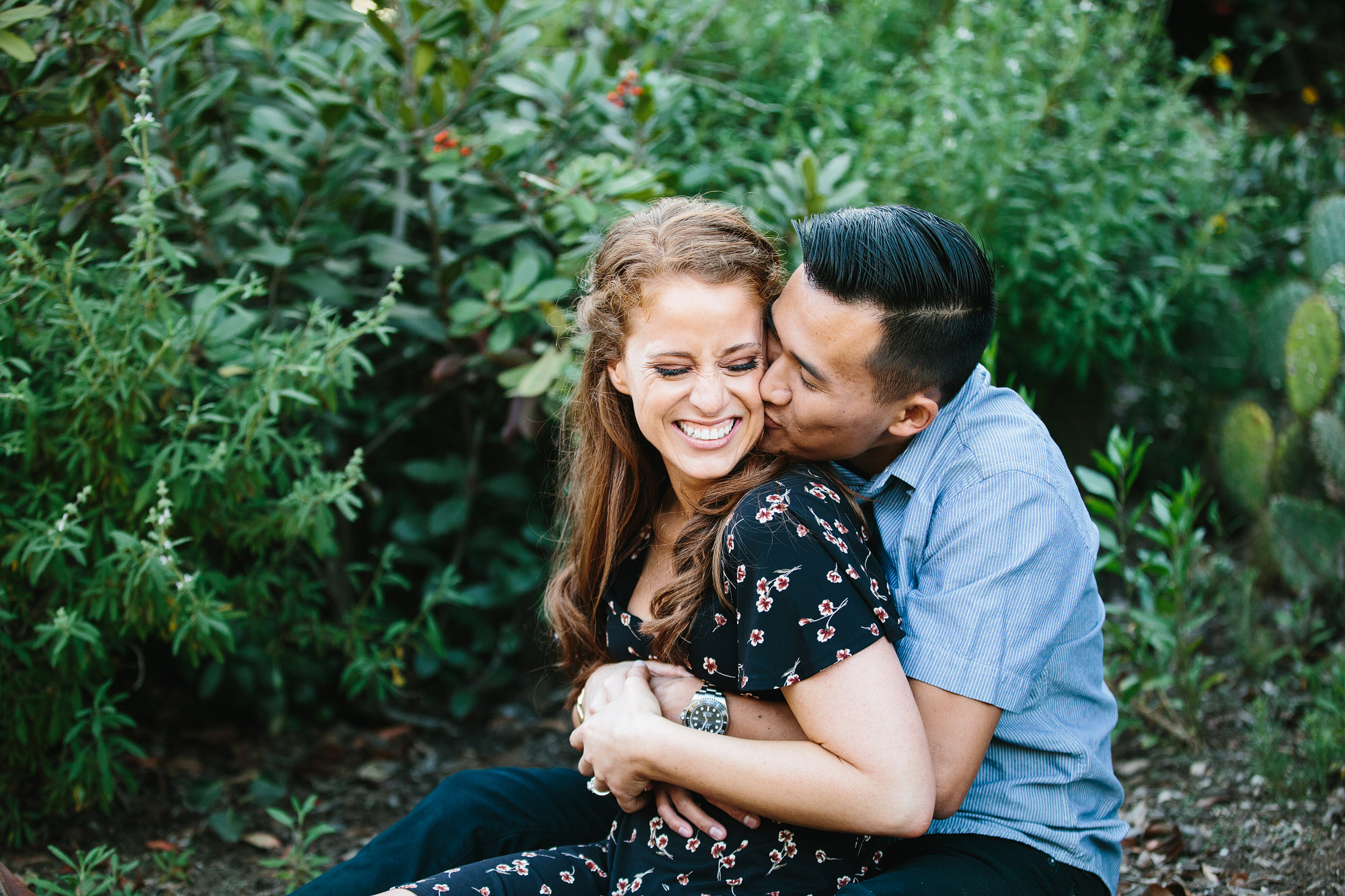 groom kissing bride to be's cheek from behind with greenery plants in background