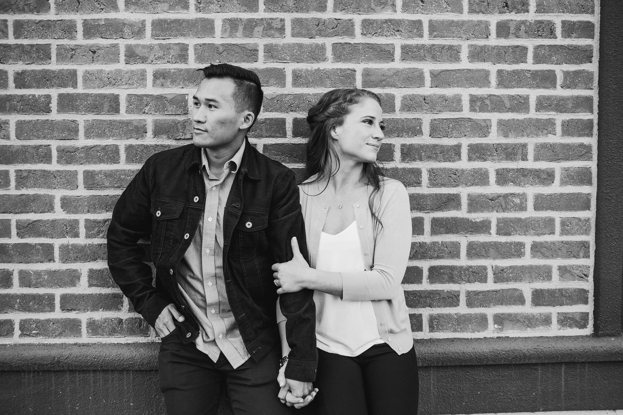 couple holding hands leaning against a brick wall in black and white