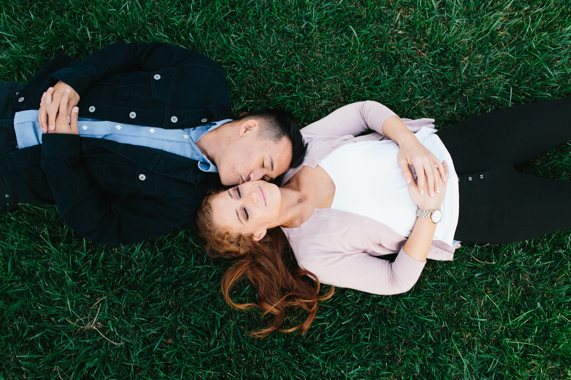 photo of young couple lying on grass guy is kissing girls cheek and she smiles